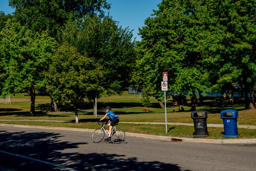 A cyclist in blue and black rides on a city street. There are road signs, trash and recycling bins, and trees.