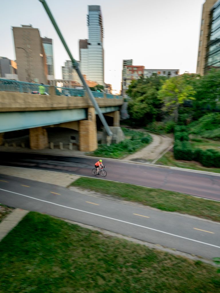 A cyclist rides on a bike path below with a bridge above and behind them. There are trees and buildings in the background.
