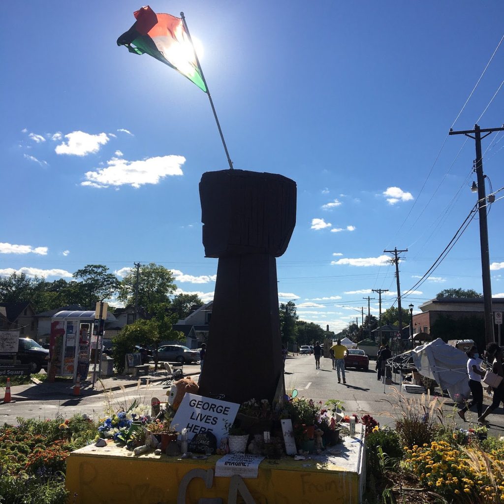 A red, black, and green protest flag flies above George Floyd Square, above a makeshift memorial, blocking the intersection of 38th and Chicago. 