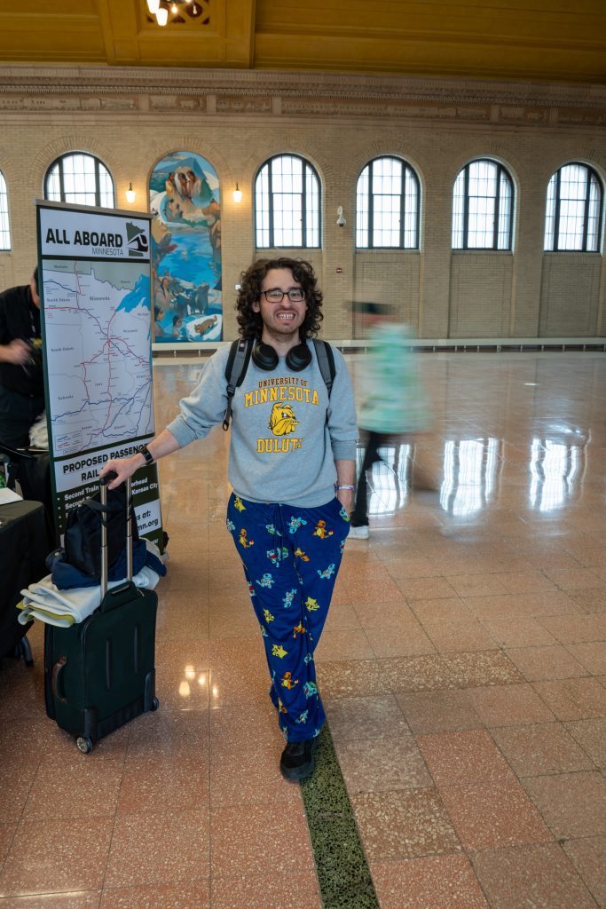 A young man standing with a rolling suitcase in a train station, smiling at the camera. 