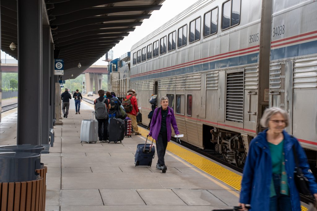An Amtrak train at a platform. Two women walk along the platform pulling luggage. In the background, people with luggage wait in line to board the train. 