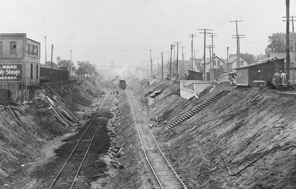 Looking west from 1st Ave of a partially constructed trench. A bridge abutment is complete on the right, and rebar for the other is in place on the left. Two uneven tracks lead to a group of workers and a steam shovel in the distance.