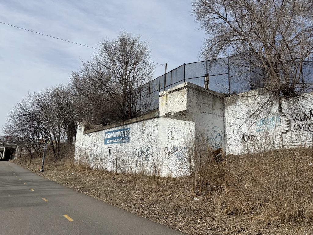 Large 3rd Avenue S bridge abutment and wing walls sit next to the Midtown Greenway covered in chipped white paint. There is a tree and other vegetation growing in and around the abutment.
