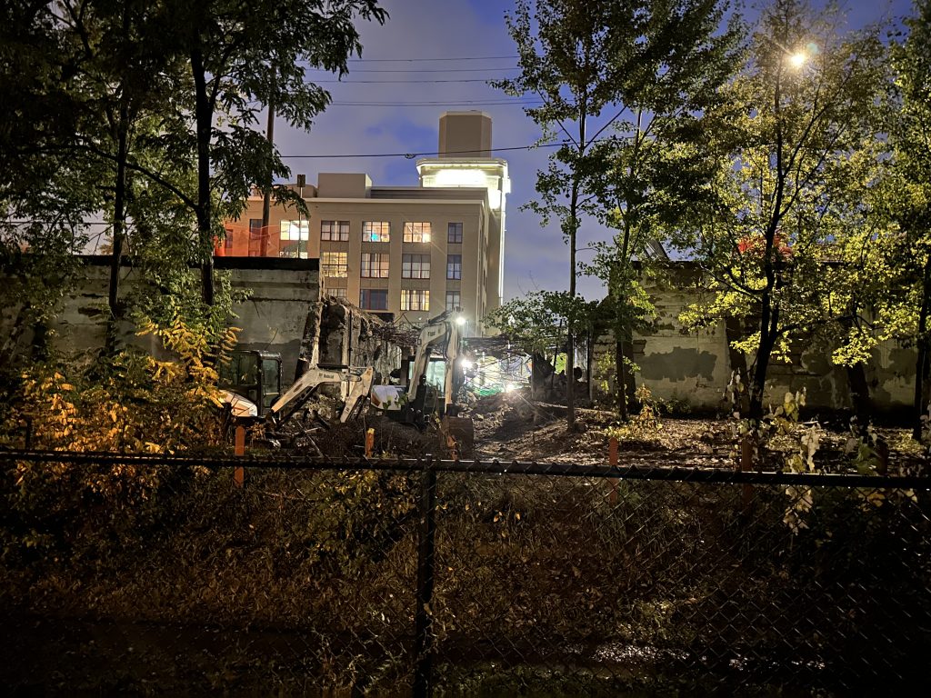 Photo at night with construction equipment parked next to a gap in the retaining wall where the bridge used to be. Rebar from the old bridge is hanging across the gap.