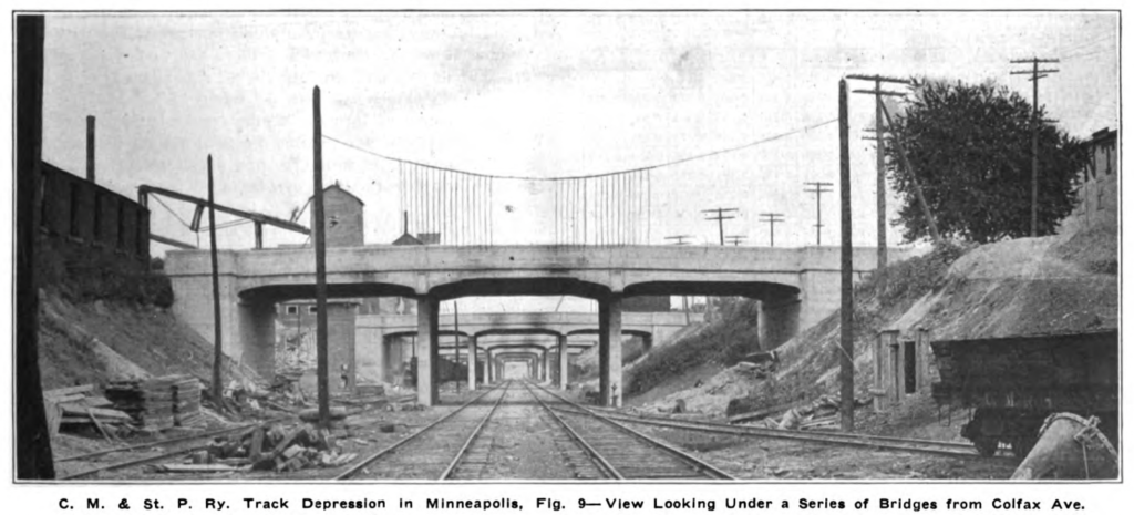 View looking through the tunnel of bridges and double track from Colfax Ave. There is equipment around and the excavation looks fresh.