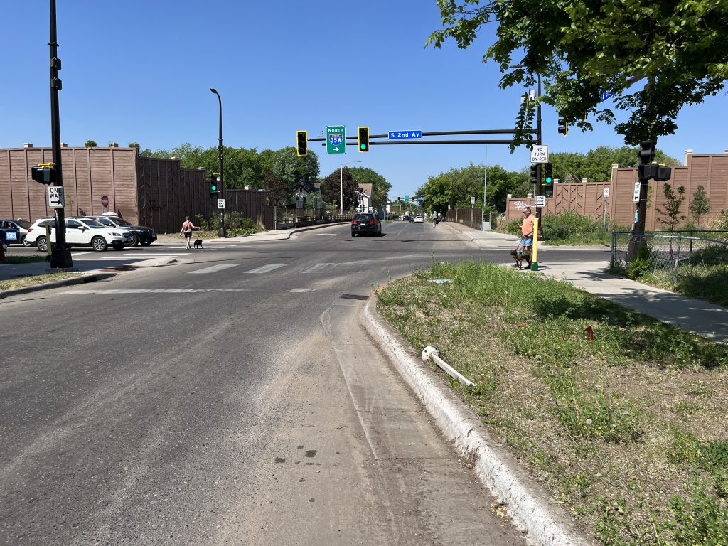 35th Street at 2nd Avenue South, with the 35th Street bridge over Interstate 35W in the background. Two pedestrians walk their dogs as traffic flows by. A white flexpost that was once part of a "bumpout" lies discarded in the tree boulevard.