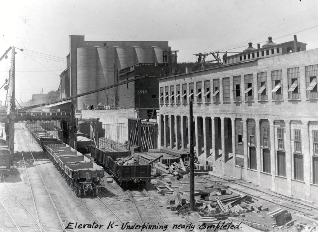 The Sheffield Elevator "K" underpinnings are nearly completed. The bridge across the trench is partially constructed. Two trains of railcars are sitting on tracks.