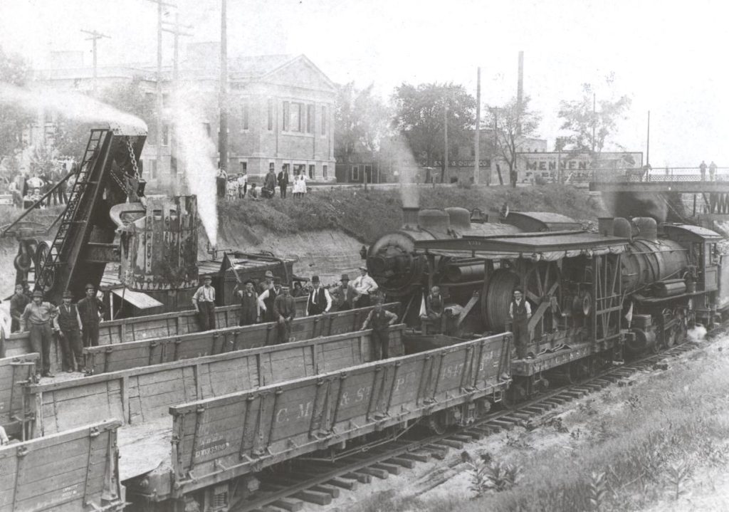 Two steam locomotives and a steam shovel are on the tracks. Connected railcars are shown empty but ready to be filled with excavated earth. The Hennepin Ave bridge over the tracks is visible in the background.