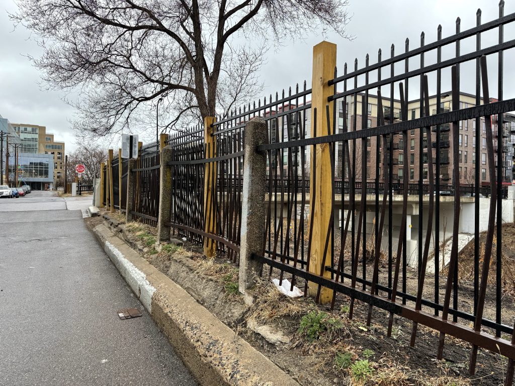 A new wood and metal fence stands a couple feet taller than the aged and historic concrete and iron fence next to it.