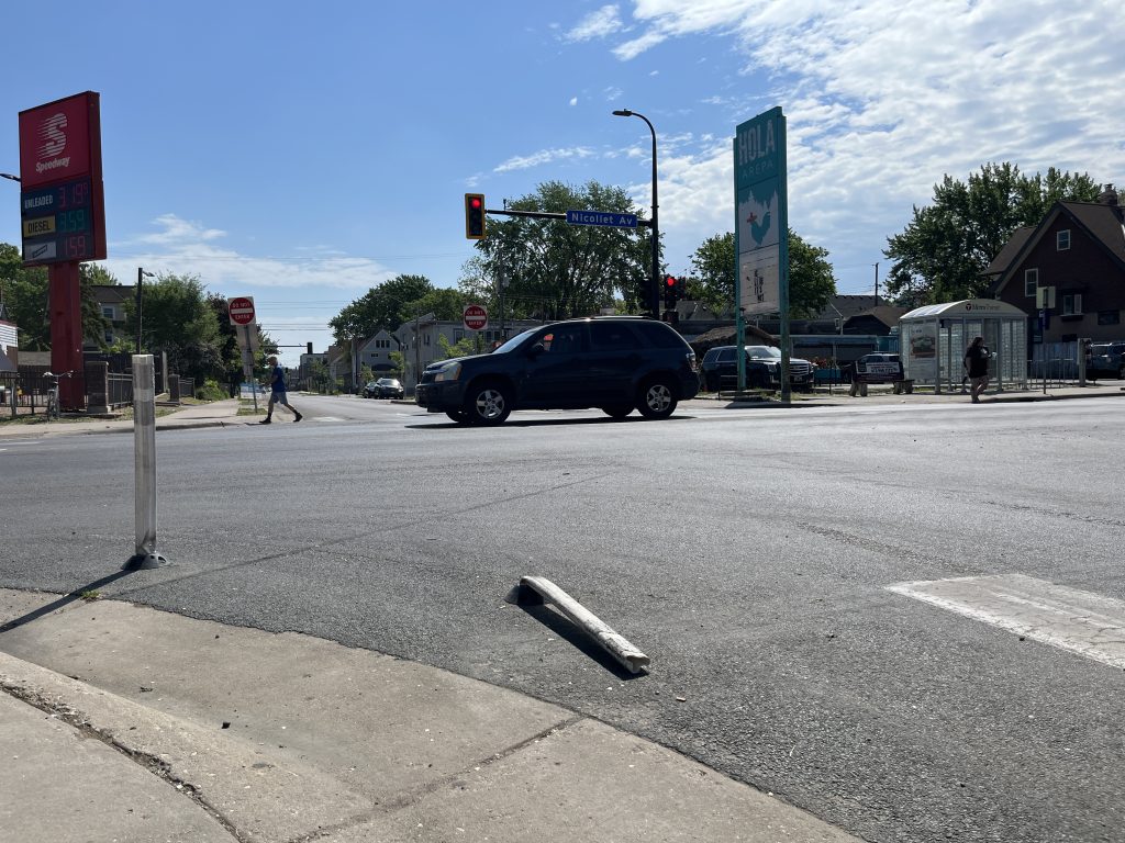 This photo is a wide view from street level of the intersection of 35th and Nicollet. A car heads north on Nicollet, passing a pedestrian near the Speedway. Hola Arepa sits in the background. There's a flattened white flexpost in the foreground.