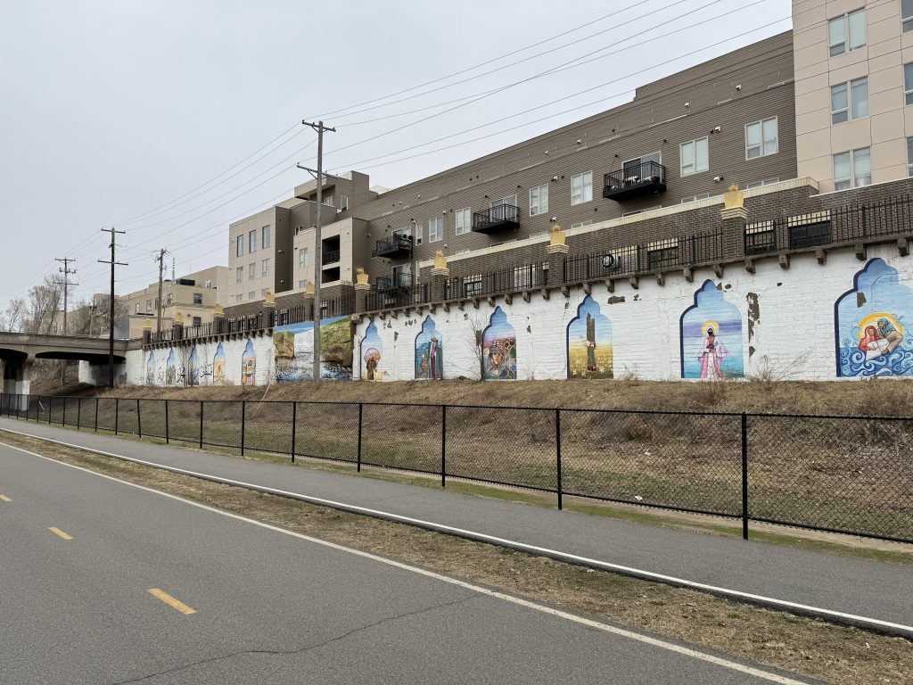White painted concrete retaining wall with colorful scenes of a mural spaced along it on the South side between Grand and Pleasant avenues.