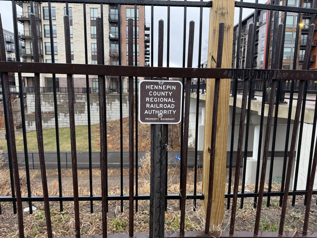 A small sign saying Hennepin County Regional Railroad Authority Property Boundary stands next to two fences at the edge of the Midtown Greenway trench.