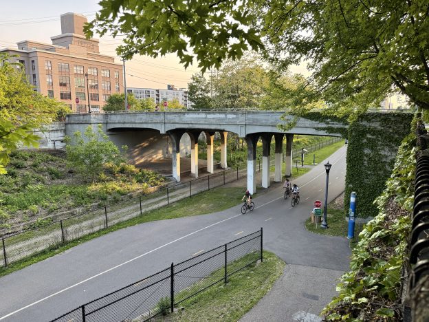 An evening sun hits the Buzza Lofts building behind the Midtown Greenway with people riding bikes under the Colfax Avenue bridge.A tree hangs over the top right of the frame.