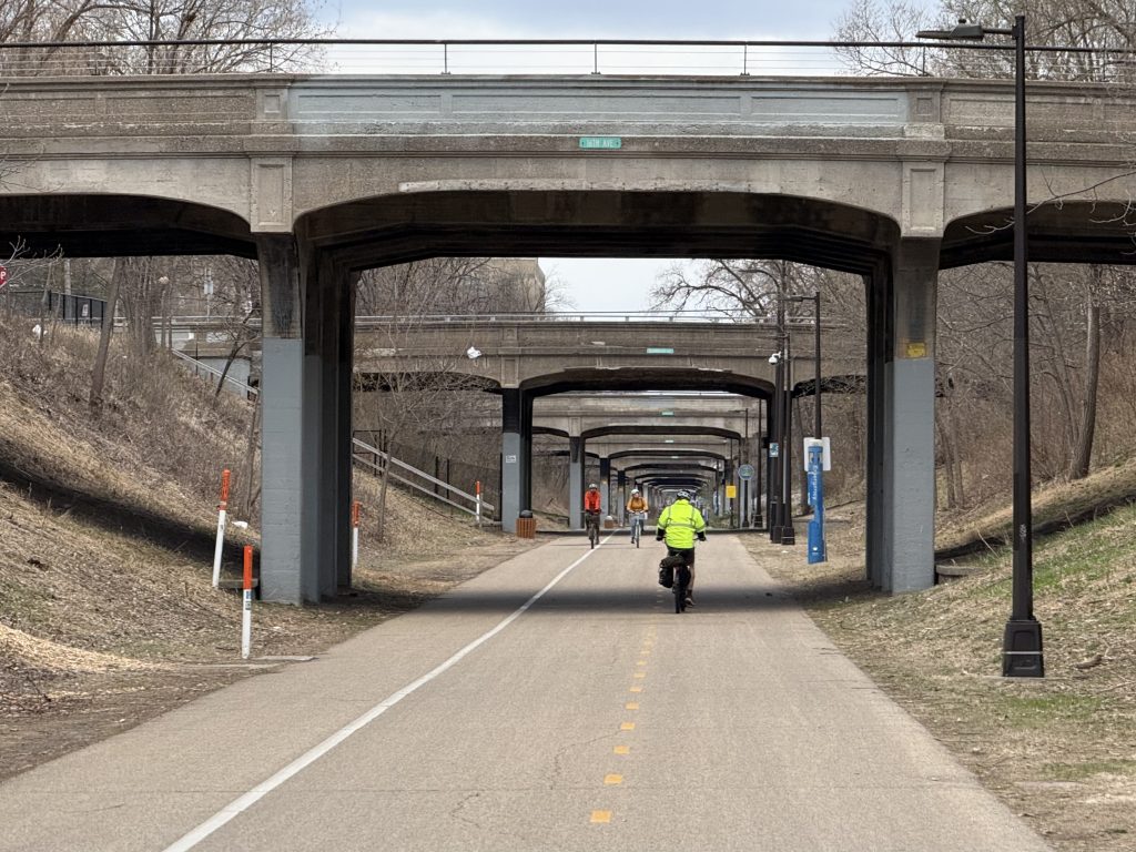 View of the Midtown Greenway looking west from 16th Ave into the tunnel of bridges with a few people biking.