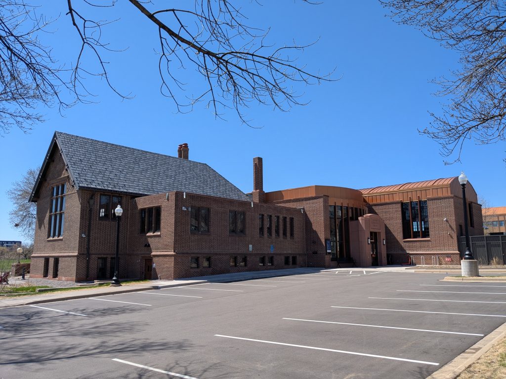 brown brick building with two-story portion at left with slate gabled roof and one story portions at right with copper flat and barrel-vaulted roofs