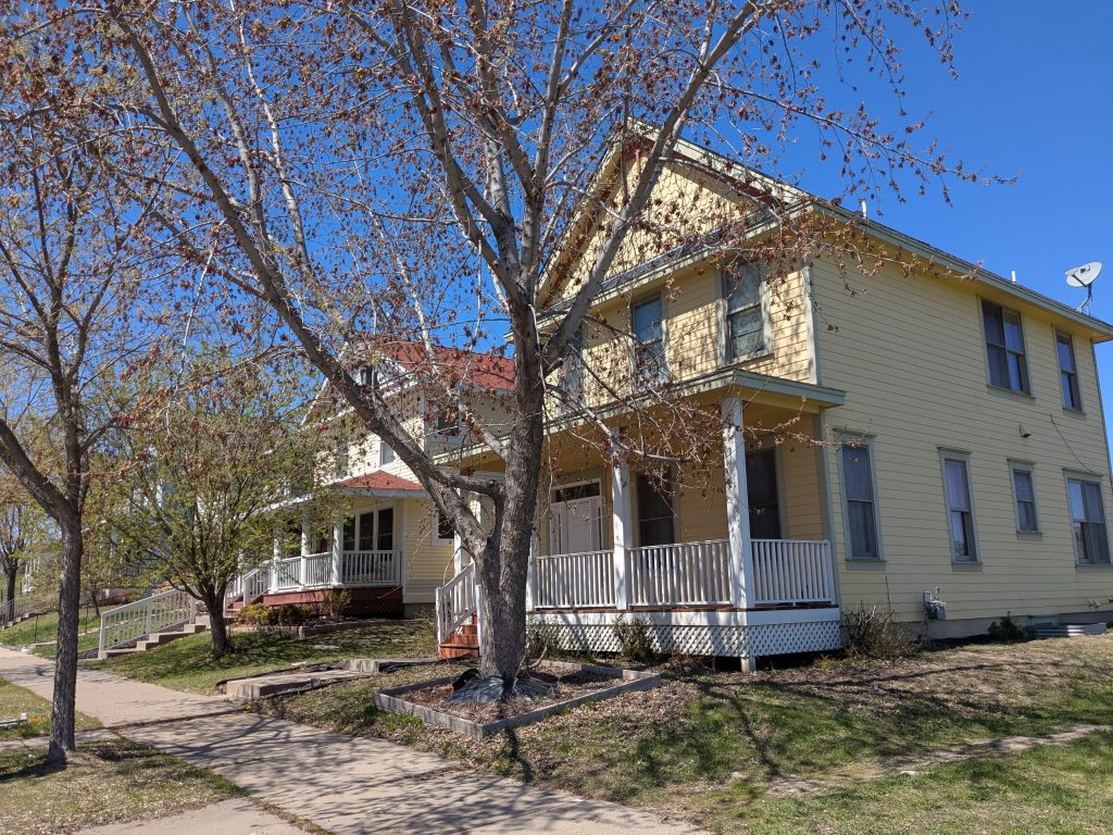 two yellow two-story greek-revival houses with porches and trees in front