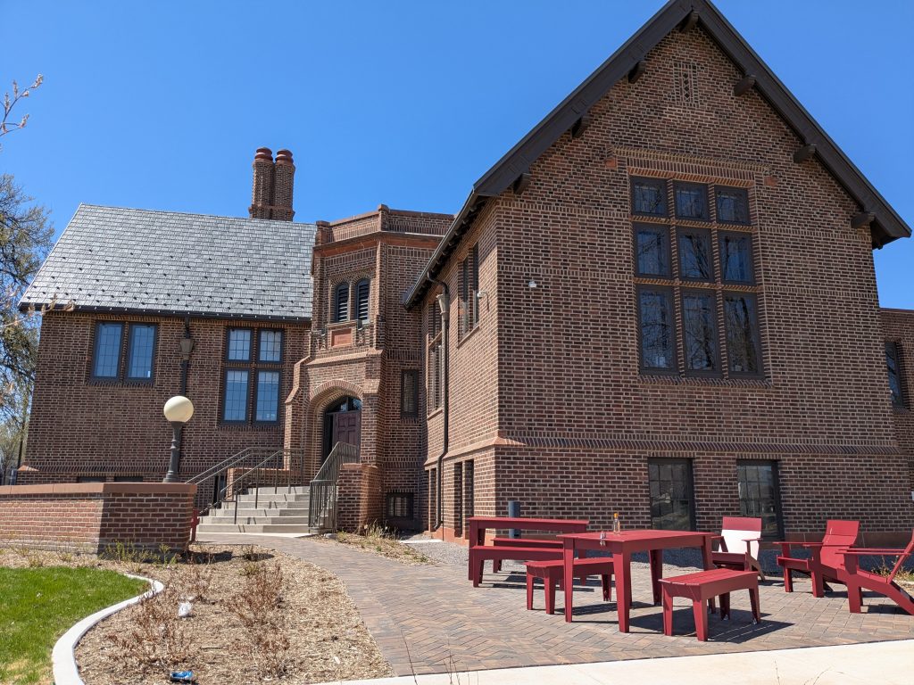 two-story brown brick building with two slate-roofed wings splayed around a turreted entryway with patio furniture in foreground
