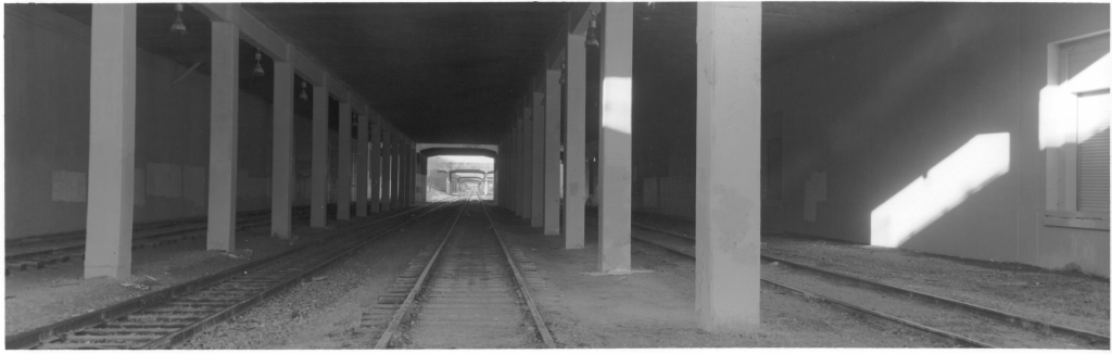A darker tunnel under the Sears warehouse with some bright sunlight shining in a couple spots too. There are four tracks visible, one on each side of the two mainline tracks.