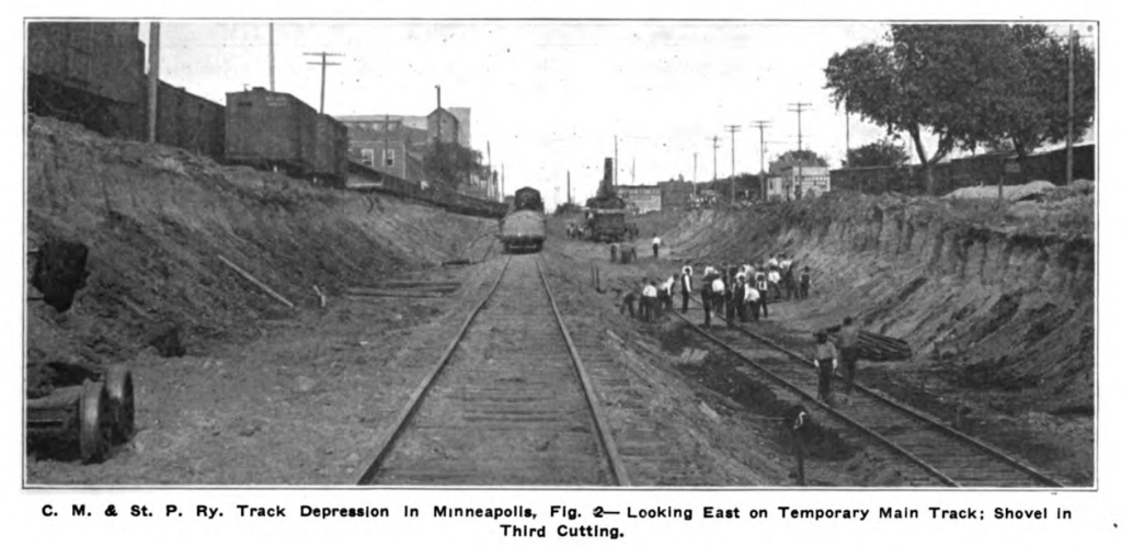 Looking east over temporary track in the trench. A dozen workers are working on the track. There are railcars above the trench on the old tracks.