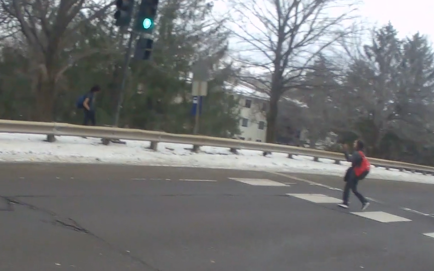 Person wearing a red backpack using a crosswalk that leads to a bus stop sign that is on the other side of a highway style barricade. Another person wearing a blue backpack and short sleeves, despite the snow on the ground, waits on the side of the barricades that is protected from the road.  