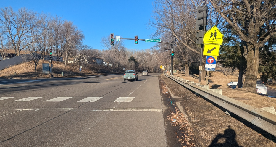 Road intersection of Cedar Lake Road and Royals Drive showing a crosswalk that leads to curb and a highway style barricade. A signpost with a crosswalk symbol and bus stop stands on the side of the barricade further from the road.  