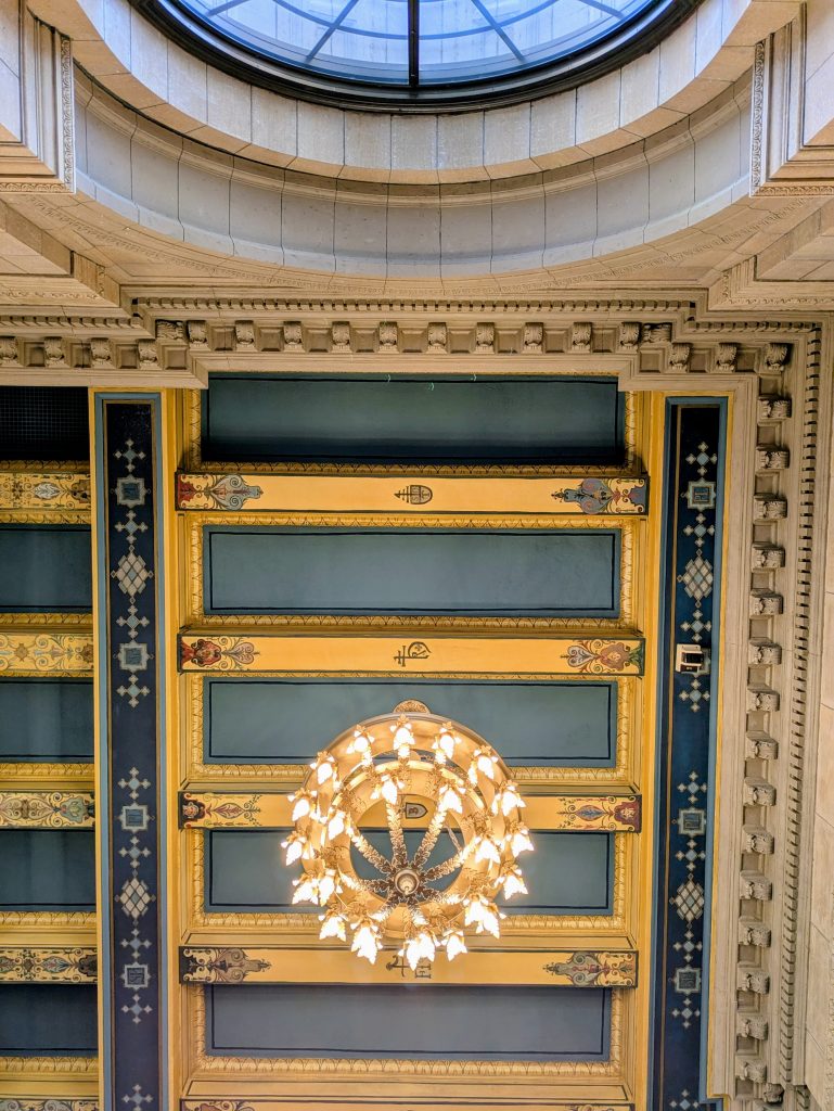 Ornate ceiling of the George Latimer Central Library reading room, featuring a large circular chandelier with glowing bulbs and intricate molding. The ceiling is decorated with gold and blue panels, floral and geometric accents, and a round skylight at the top.