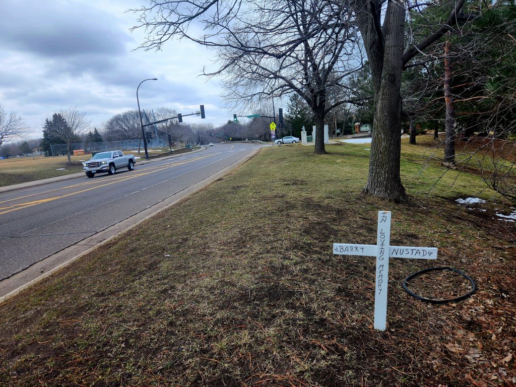 Cross bearing the text "Barry Nustad, In Loving Memory" situated next to Cedar Lake Road.