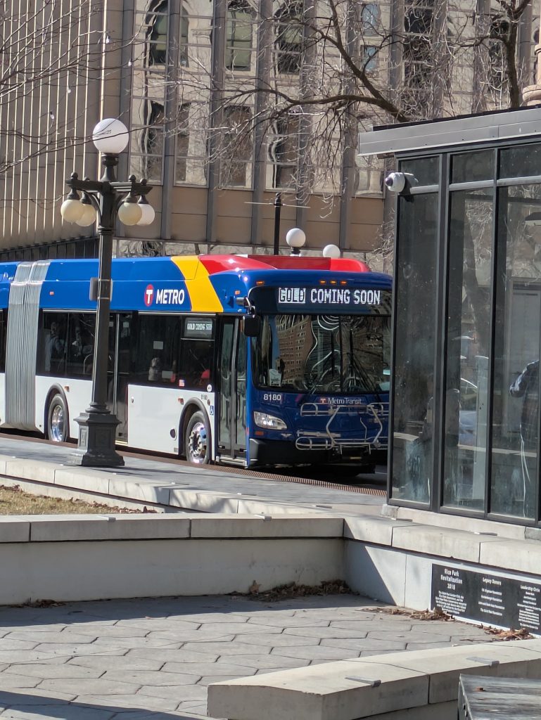 Metro Transit bus with "GOLD COMING SOON" displayed on its sign, parked near a city sidewalk.