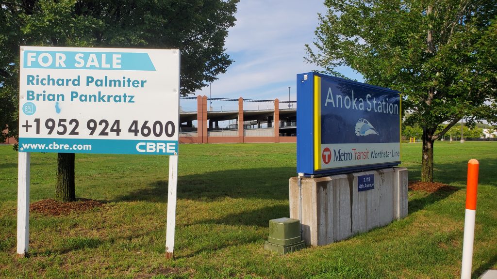 A for sale sign adjacent to the sign for Anoka Station, with the station in the background.