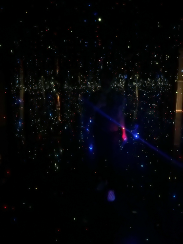 A photograph of the author in Yayoi Kusama’s Infinity Mirror Room. The room is dark, the walls are covered in mirrors, and tiny points of light look like stars surrounding the viewer like fireflies. If you close your eyes, it feels like a passing smell of cherry trees in spring.
