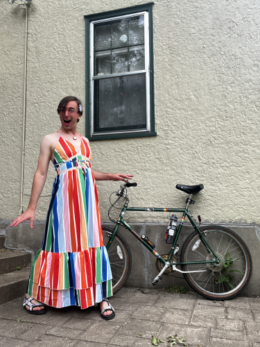A jubilant woman poses in a rainbow dress in front of a bike.