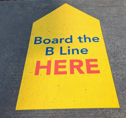 A stylized yellow arrow on the sidewalk reads "Board the B Line HERE."