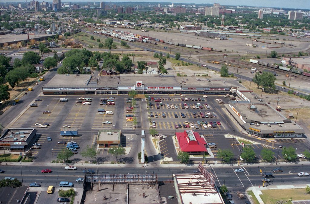 Looking North from Lake Street with a strip mall in the foreground and with Hiawatha Avenue and the rail yard in the background. Amidst the rail storage, there is a massive parking lot.