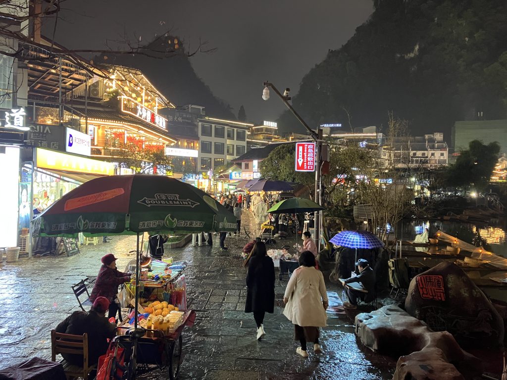 Nighttime view of same cobblestone street as previous photo with many people walking and filled with venders with umbrella covered carts.