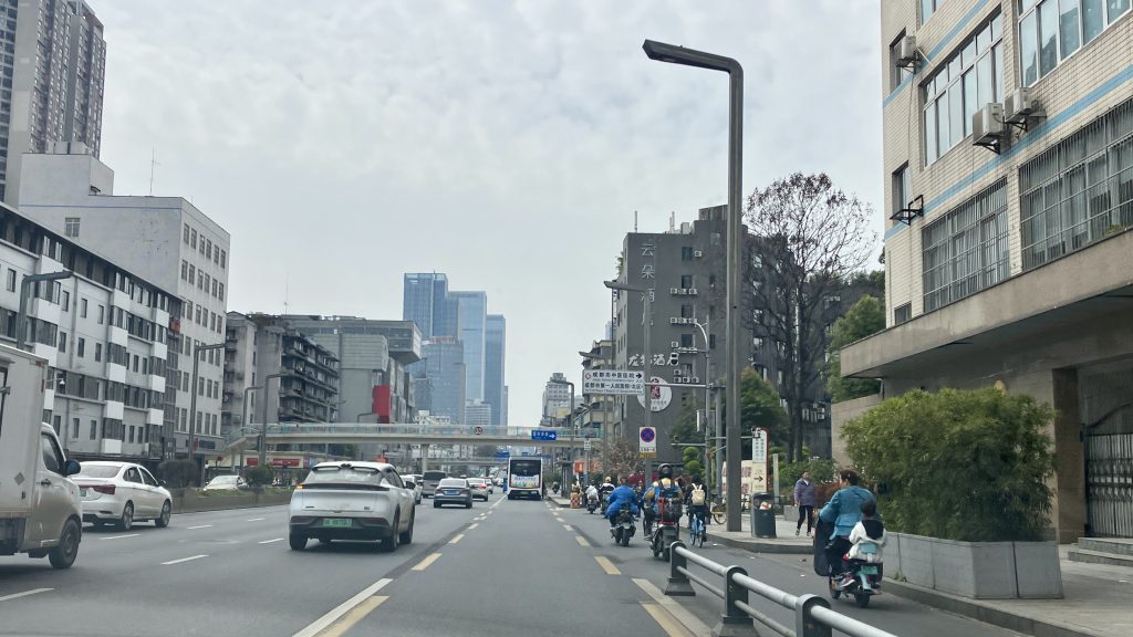 A wide streetscape in Chengdu with  vehicle lanes, bike lanes and sidewalks, lined with greyscale towers and skyscrapers
