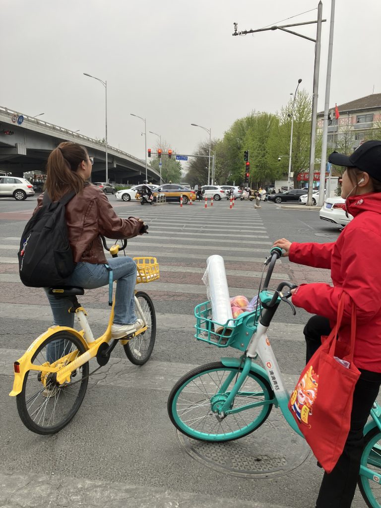 Two people on Chinese bike share bikes wait to cross large intersection  with refuge island ahead, midway of the crossing.