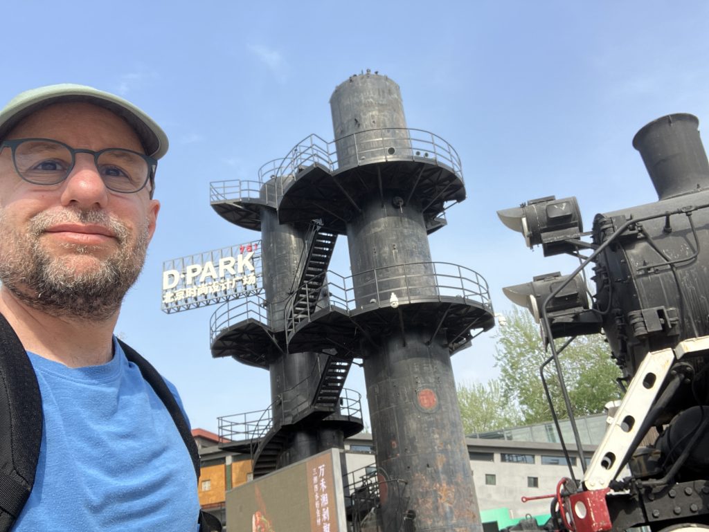 Author selfie in front of large, old industrial smokestacks and diesel locomotive train engine.