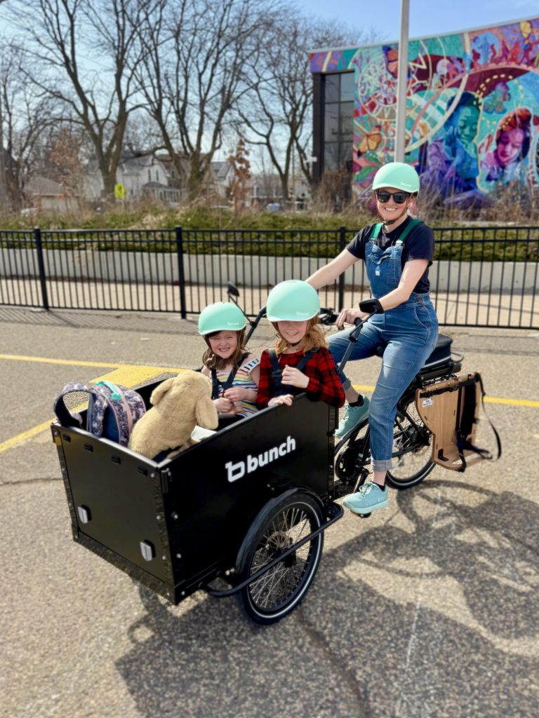 Laura smiles at the camera from the seat of a front-loading cargo bike that contains two smiling children, a backpack, and a large plushie. Laura and her kids have matching mint green helmets.