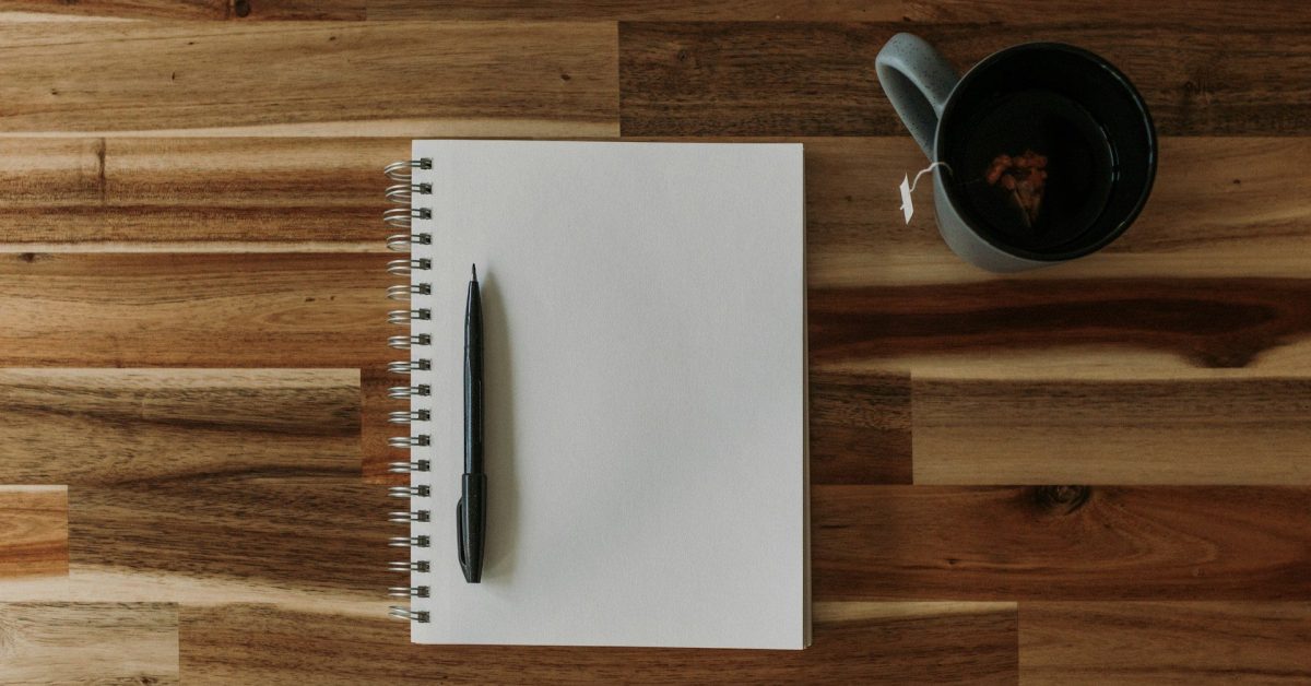 A pen rests on a blank page of a spiral notebook with a cup of tea nearby.