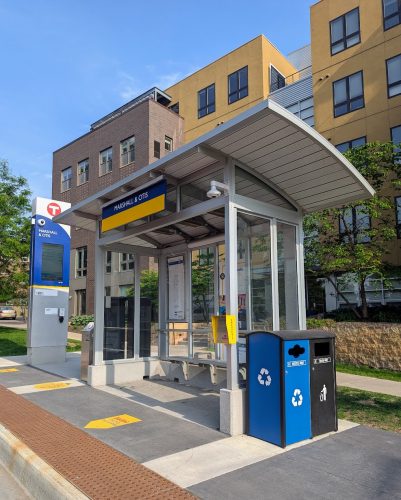 A bus stop with a shelter, trash and recycling bins and an electronic arrivals sign.