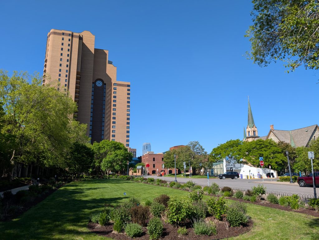 Green landscaping with highrise building in the background featuring a tall arched inset with a rose window at the top; a church steeple is off to the side.