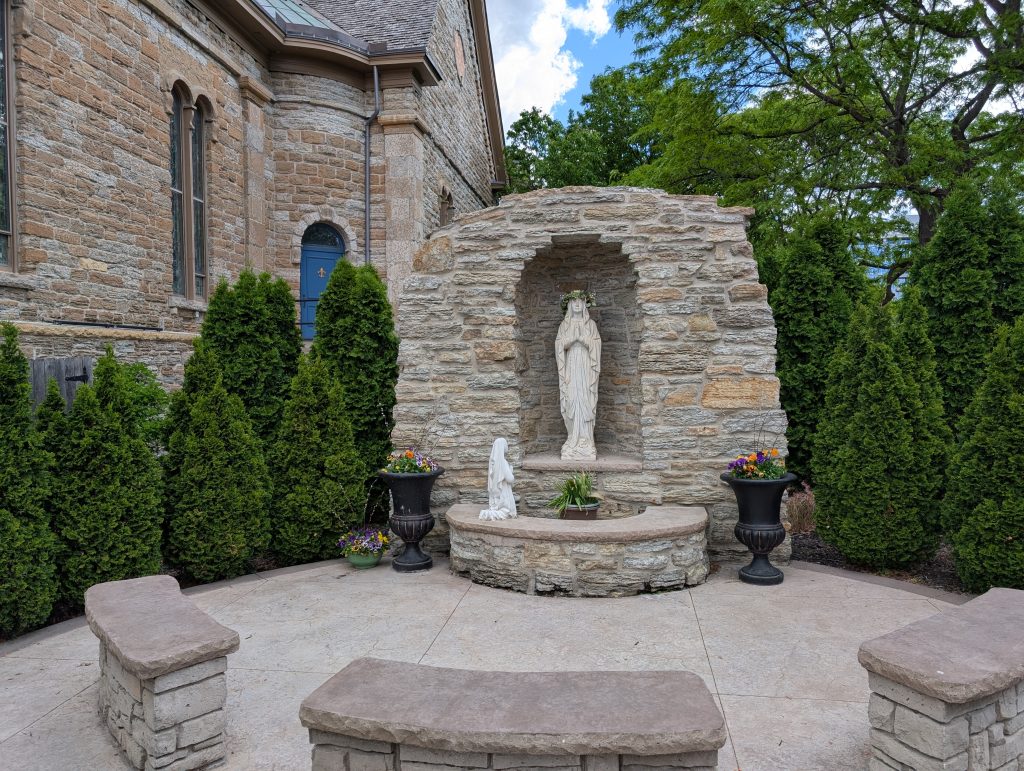 Stone grotto containing a white statue; a smaller white statue kneels in front  on a bench; additional benches are further away.