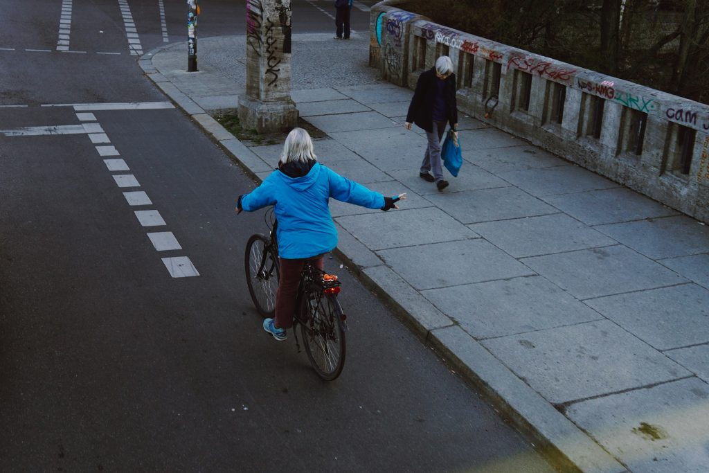 A gray-haired woman on a bicycle in the city signals a right-hand turn as she crosses a bridge.