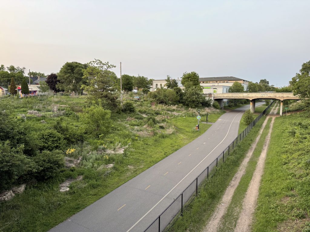 Overlooking the Midtown Greenway with an overgrown SOO Line garden to the left of the paths and the Harriet Avenue bridge in the background.