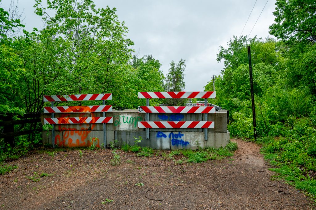 Two red and white hazard signs block a road surrounded by greenery and an unpaved trail