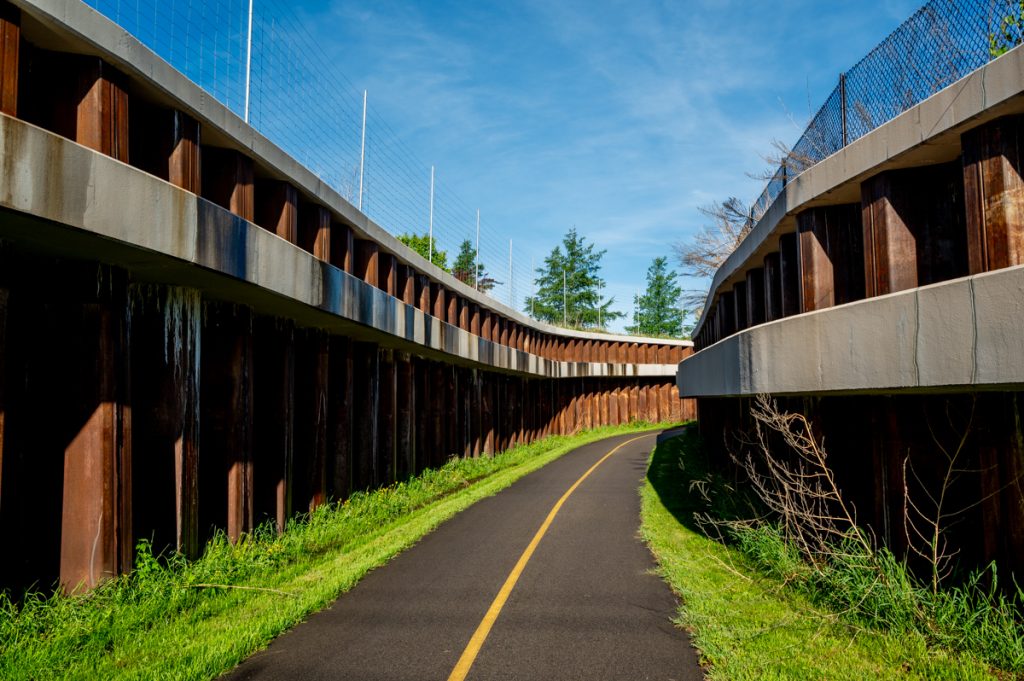 A paved Regional Trail surrounded by high metal barriers leading to the Minnesota Landscape Arboretum
