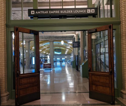 Wooden doors open onto the marble halls of Union Depot, with a sign overhead pointing toward the Amtrak Empire Builder Lounge.