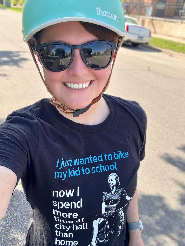 Selfie of Laura wearing a shirt designed by Tom Flood that reads “I just wanted to bike my kid to school now I spend more time at city hall than home.” w/ a photo of a woman on a bike.