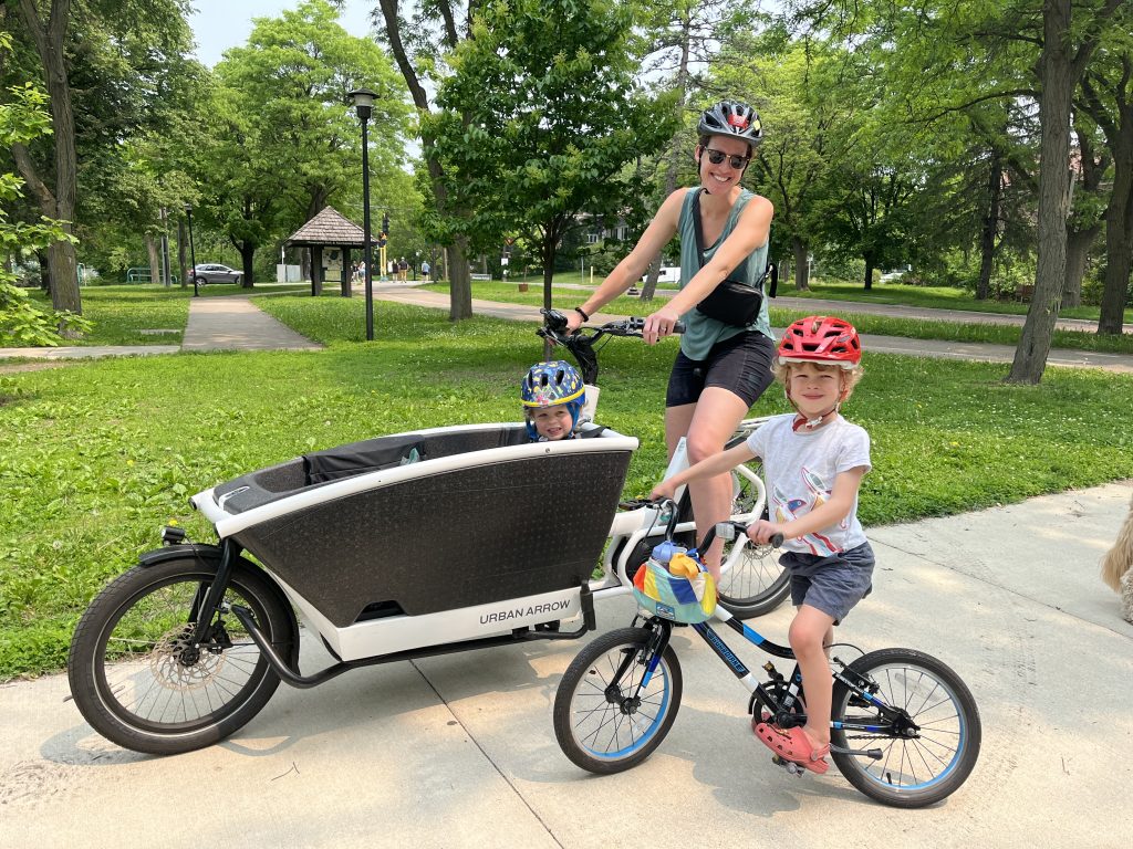 A woman sits astride a cargo bike that has a child in the front holder next to a child on their own small bike.