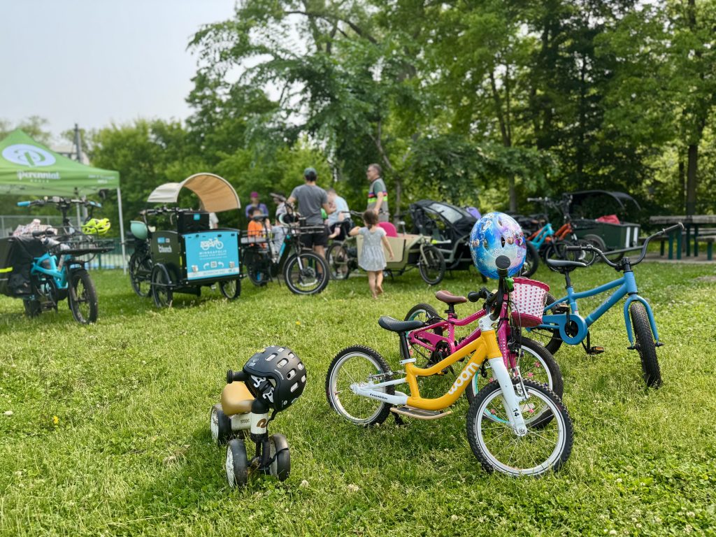 Small children's bikes sit on the grass in front of adult cargo bikes at the show & tell event.